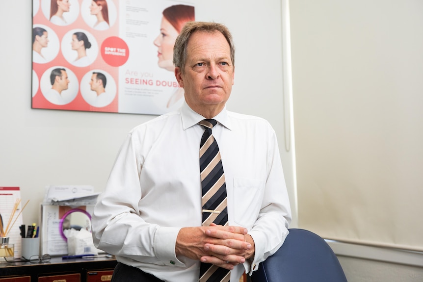 A man in a white shirt with a black and gold tie leans on a medical examination chair.