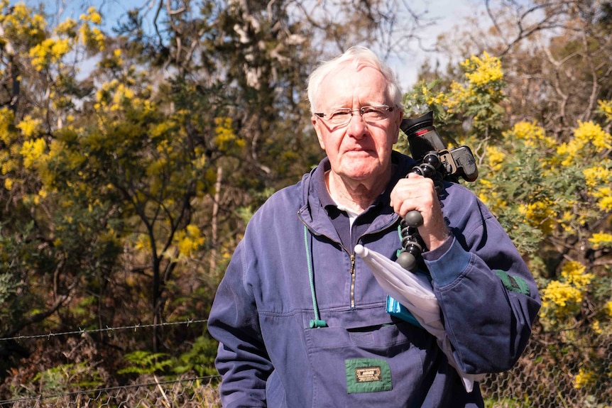 Picture of a man in a blue jumper holding a camera with trees in the background.