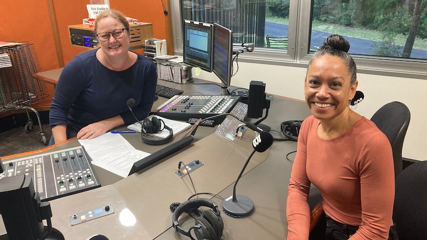 Two women sit opposite each other with a radio studio panel between them