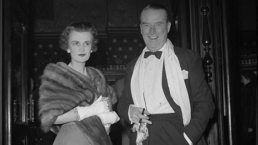 Margaret Campbell dressed in a fur coat clutches a purse standing next to a man dressed in a suit with a bowtie.