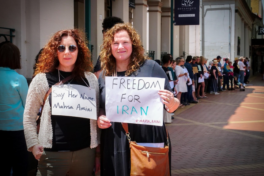 Two women hold placards protesting violence in Iran in front of a long line of people holding similar signs