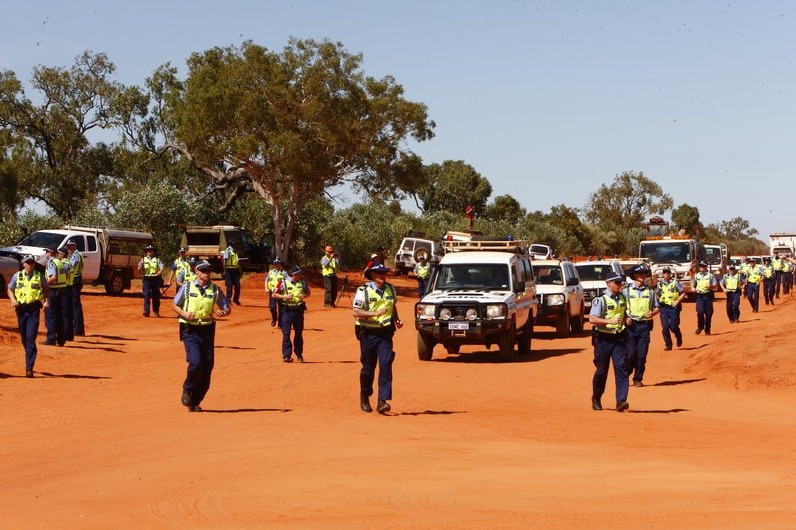 A group of police run alongside a line of vehicles on a red dirt road.