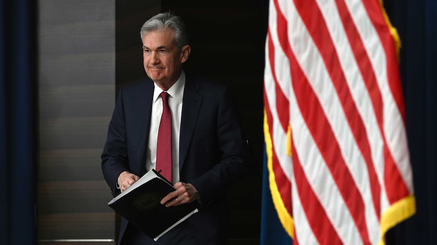 US Federal Reserve chair Jerome Powell walks with a clipboard in behind an American flag.