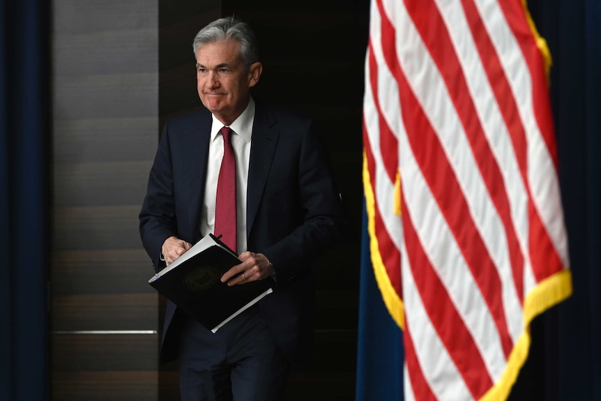 US Federal Reserve chair Jerome Powell walks with a clipboard in behind an American flag.