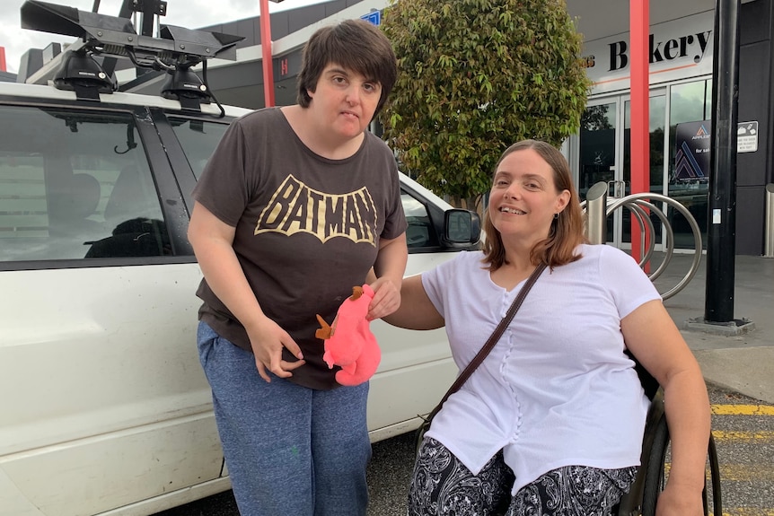 A woman seated in a wheelchair with her arm around a younger woman who is standing next to a car.