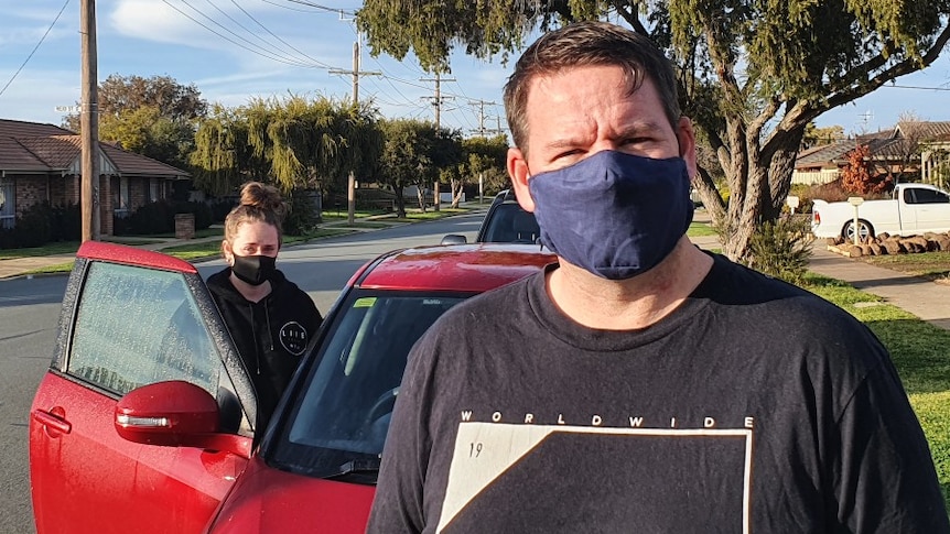 A man wearing a mask standing in front of a red car with a young girl standing near the drivers door of the car