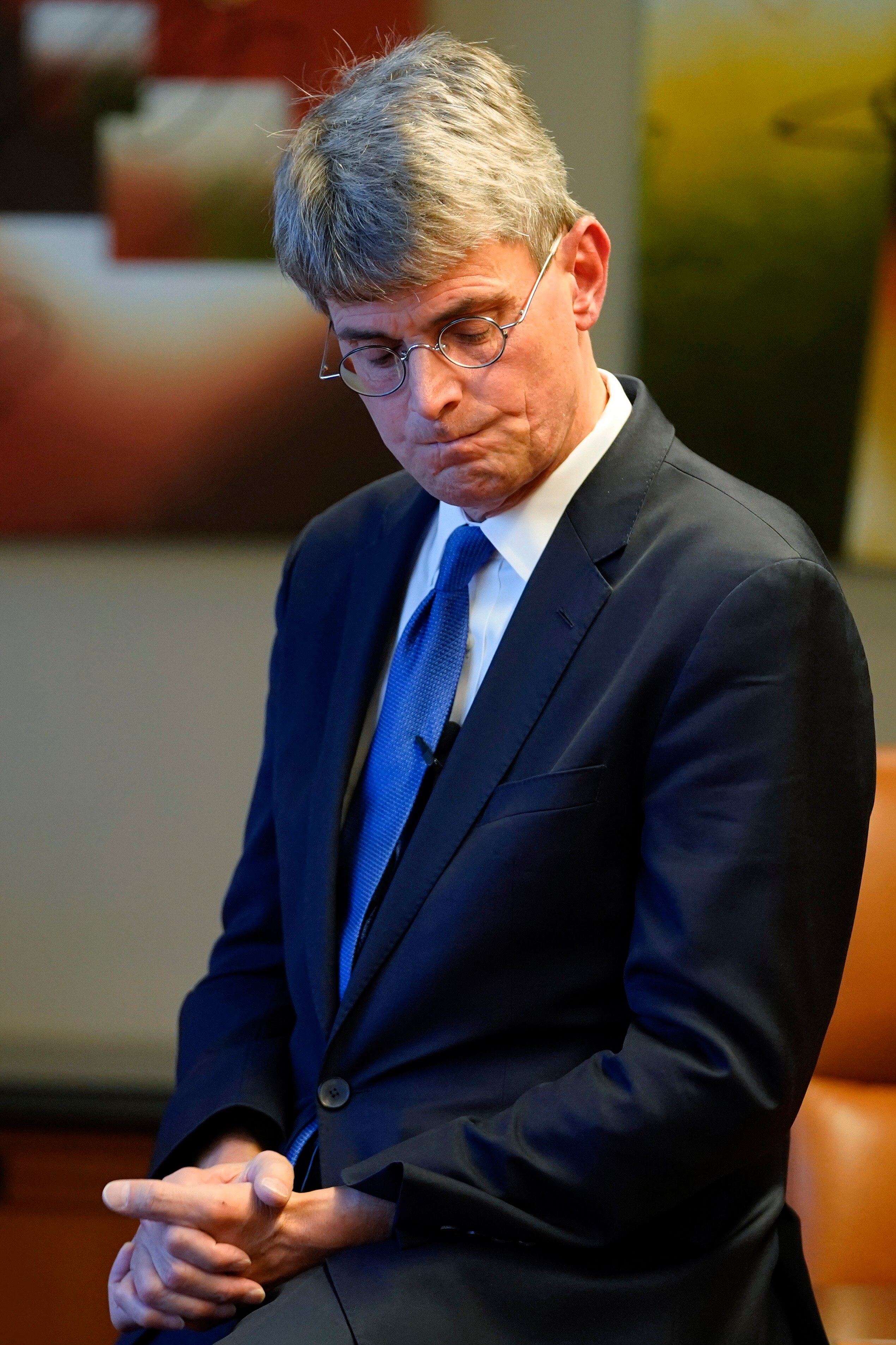 Man in a suit with a blue tie grimaces as he looks downward. 