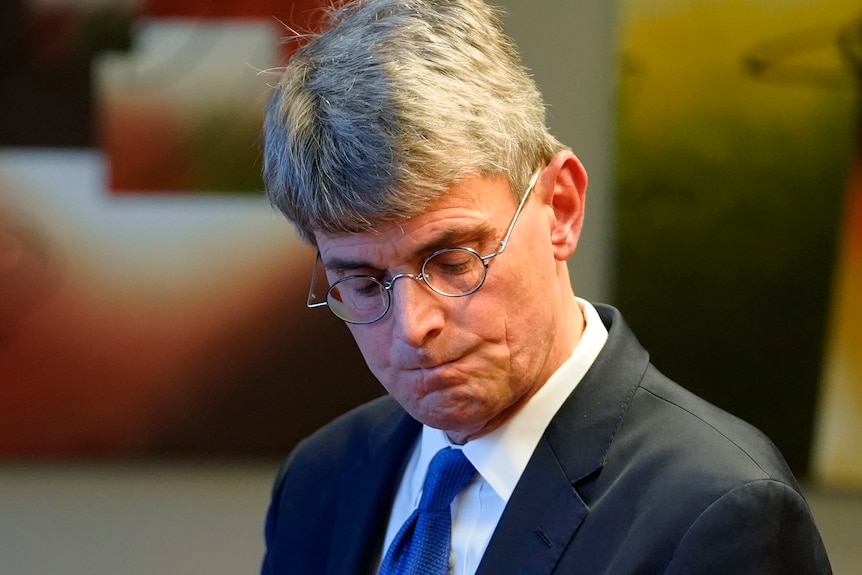 Man in a suit with a blue tie grimaces as he looks downward. 