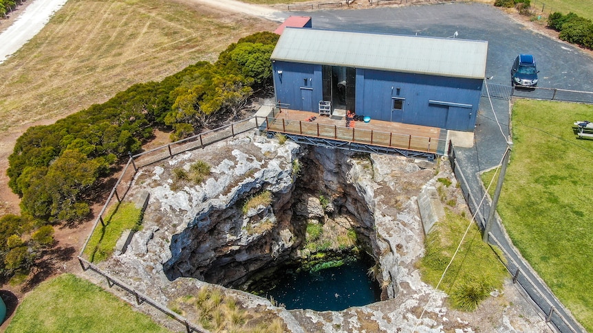 A large shed sits on the edge of a large sinkhole, a natural hole in the ground made of limestone walls.