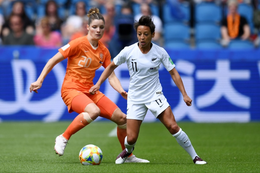 Two soccer players, one in orange and one in white, look down at a ball in the grass