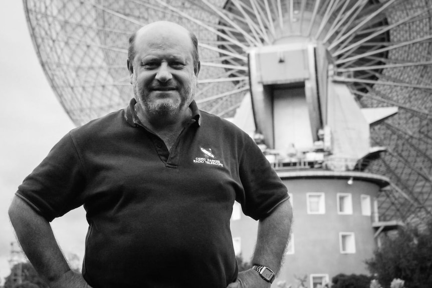 A black and white photo of a man standing in front of a large radio telescope.