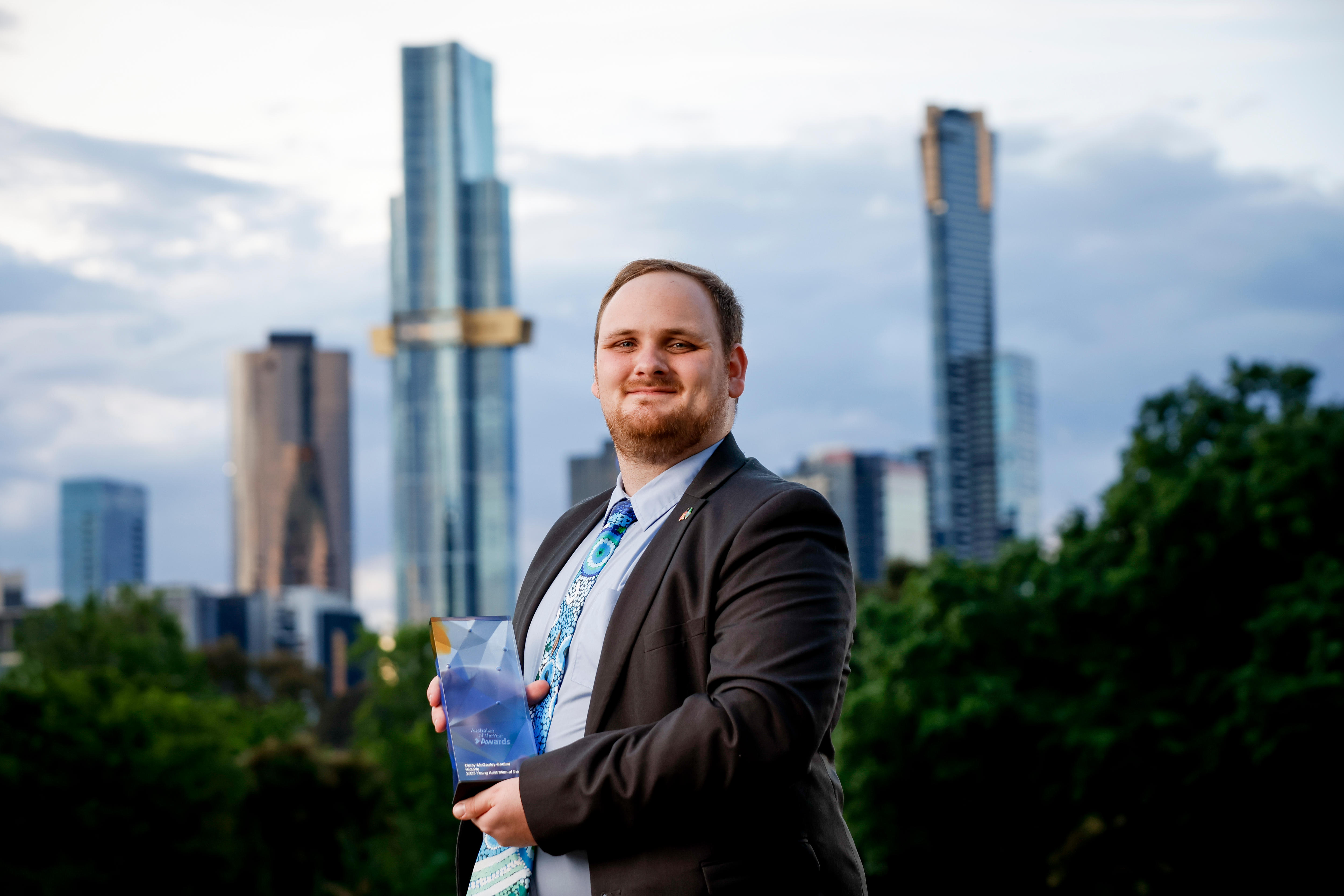 A young man standing in front of city building holding his australian of the year award