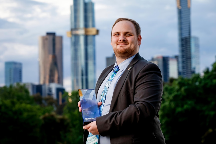 A young man standing in front of city building holding his australian of the year award