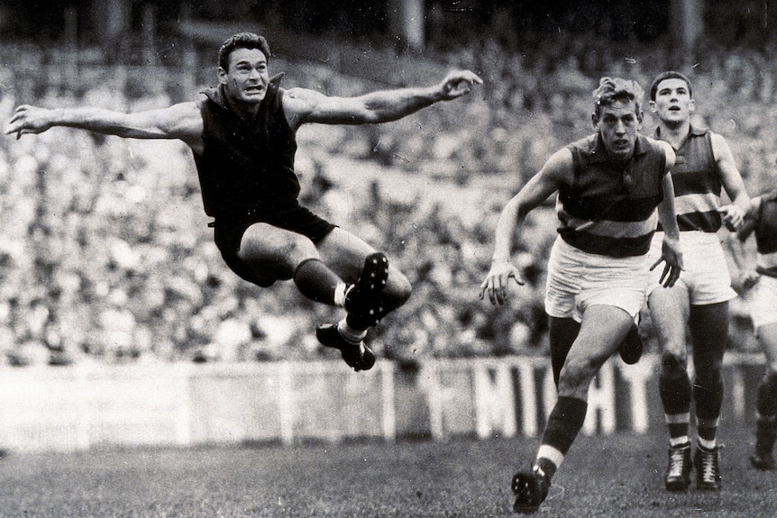 Ron Barassi is airborne as he kicks the ball with both arms stretched wide