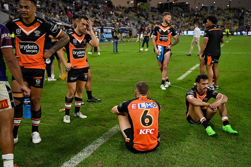 A group of Wests Tigers stand around looking dejected or sit on the ground with their hands on their knees.