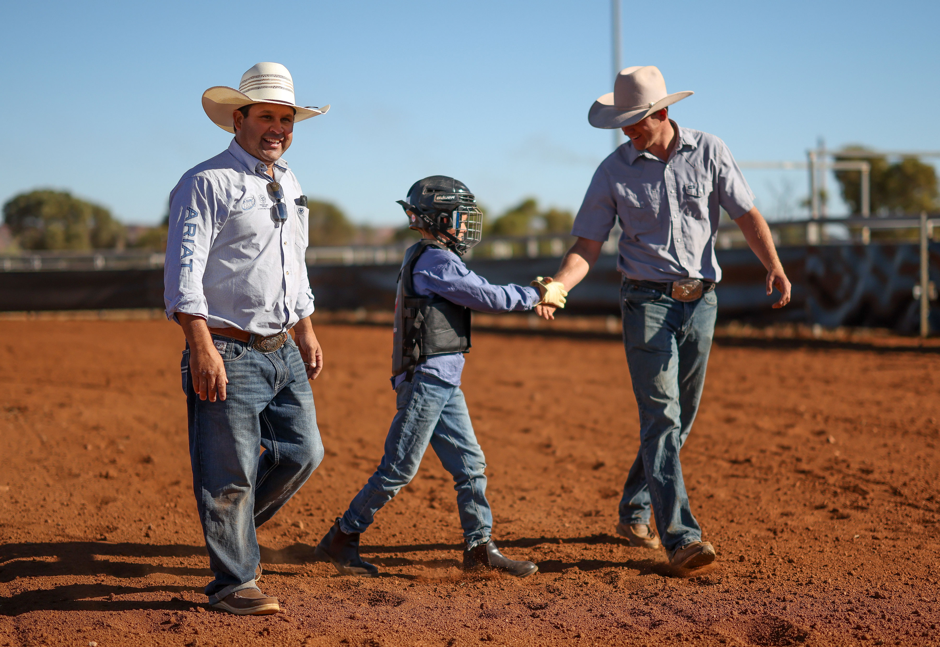 A young rider wearing a helmet and protective vest shakes hands with an adult instructor in a red dirt rodeo arena.