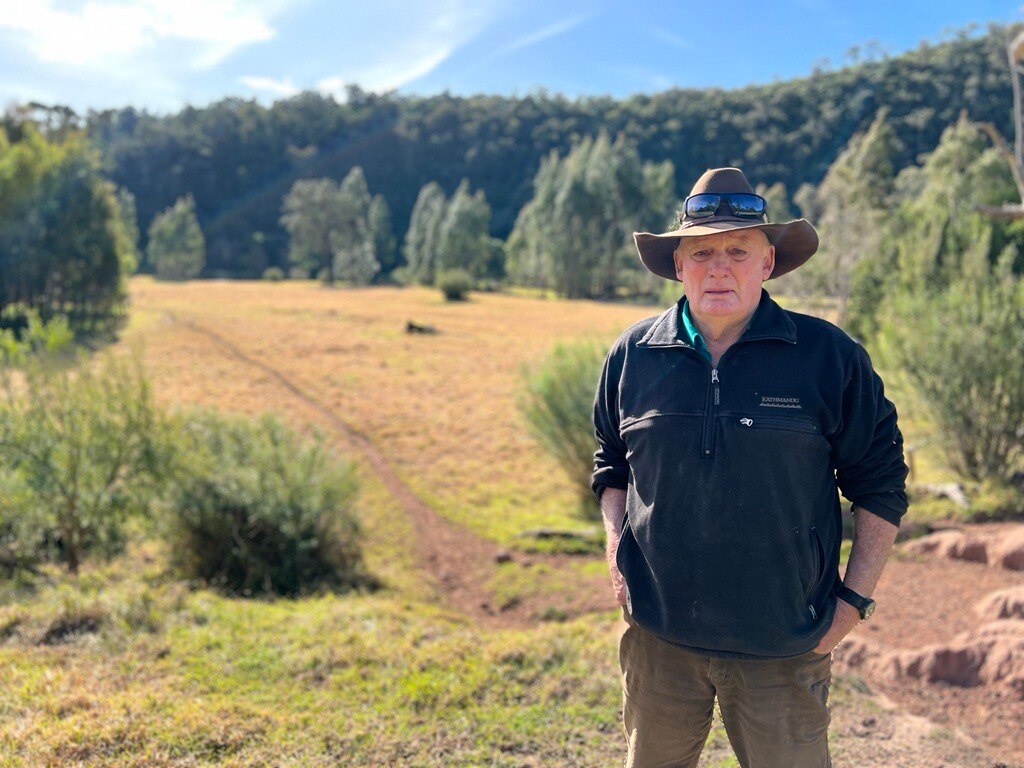 Trevor stands on a ridge above a paddock leading down to the river.  He wears a broad hat and a dark jumper.