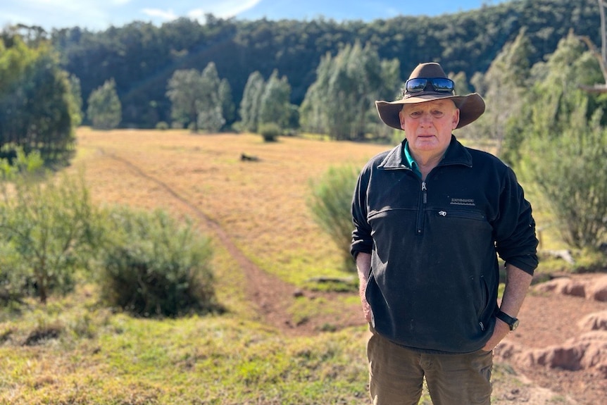 Trevor stands on a ridge above a paddock leading down to the river.  He wears a broad hat and a dark jumper.