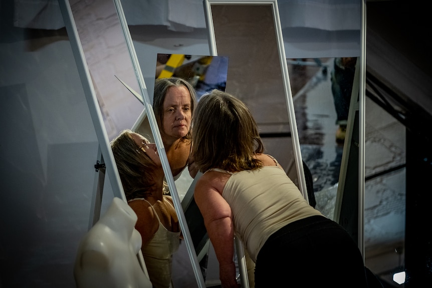 Leysa leans over to look into the mirror and is reflected in other mirrors in the surrounding space.