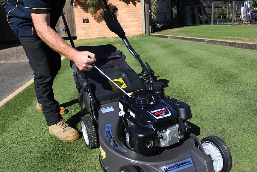 A man in workwear and boots pulling the rip cord of a black four-stroke lawn mower