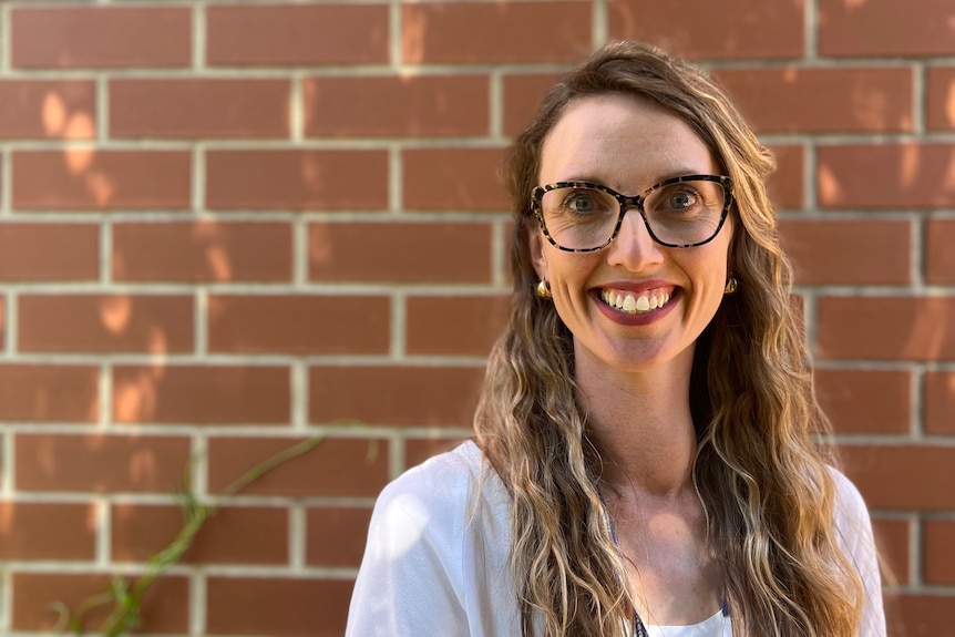 Woman with curly blonde hair and wide-brimmed reading glasses smiles at camera in front of red brick wall 