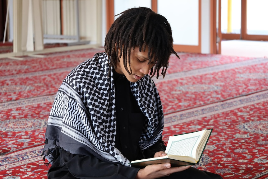 A young man sits on a red patterned carpet reading the koran