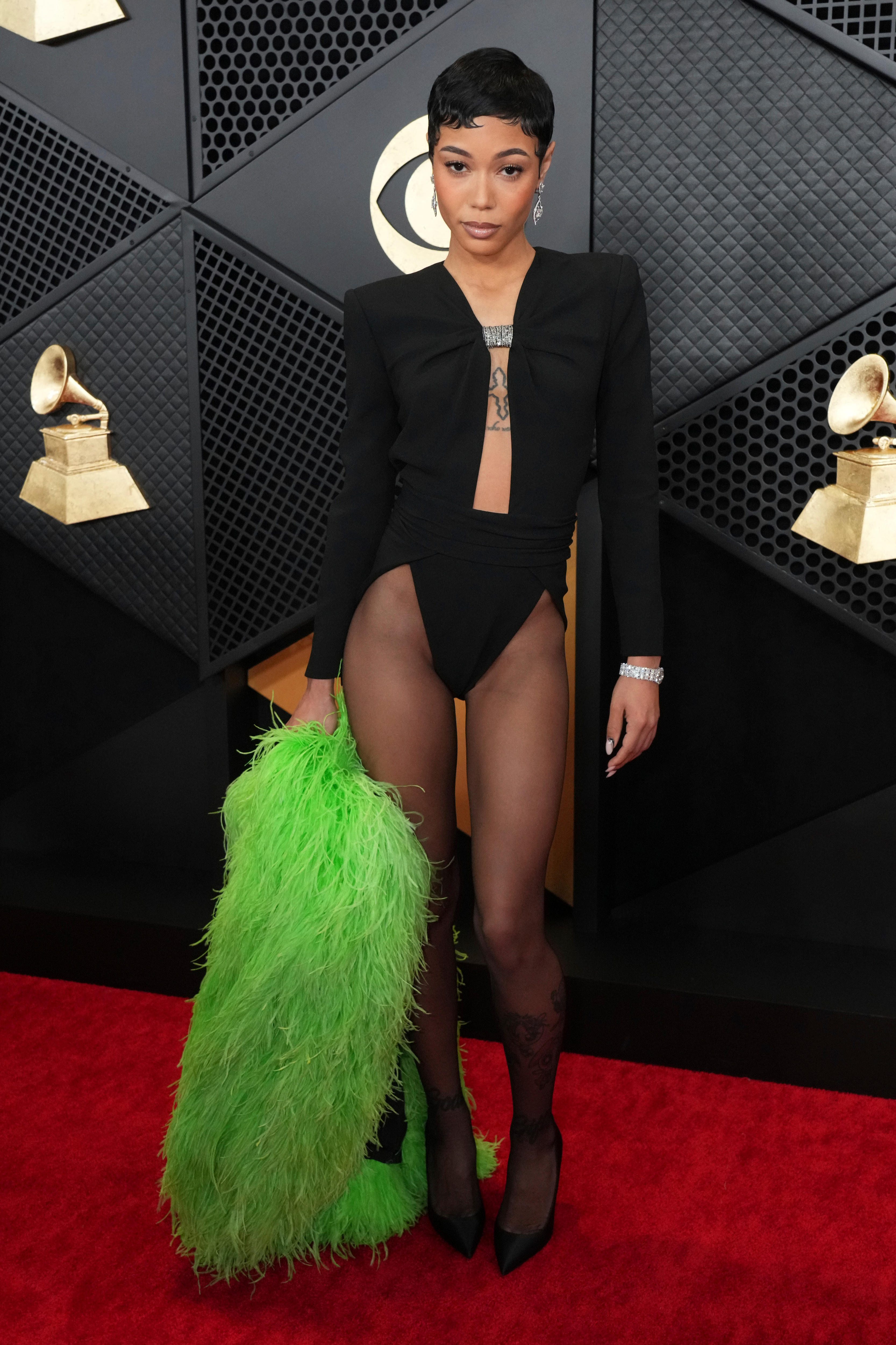 Coi Leray wearing a balack blazer-like leotard with stockings and carrying a fluffy green jacket