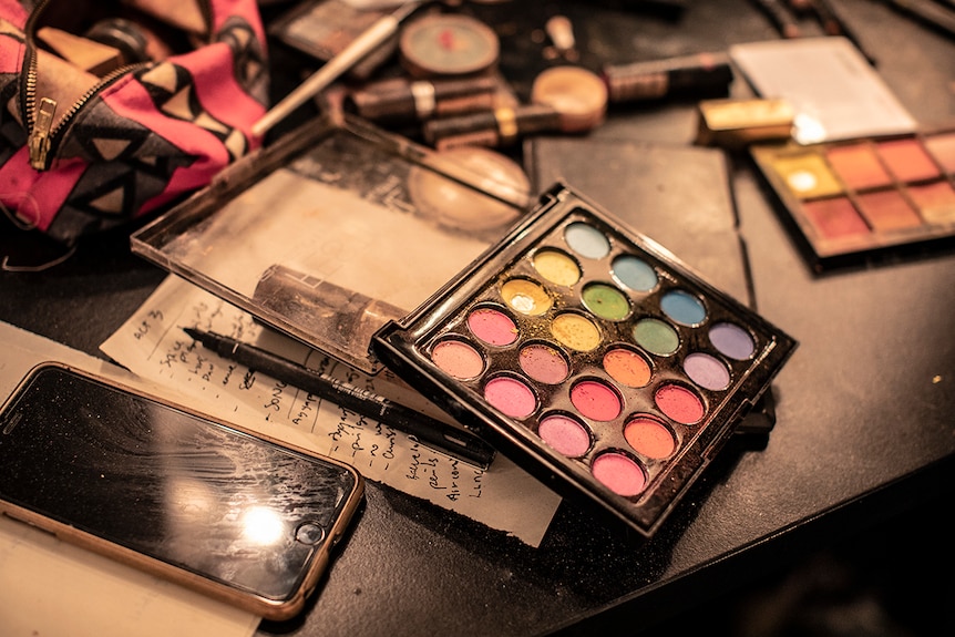 Close-up photo of make-up eye shadow palette on dressing room table top next to iPhone, pen and paper notes.