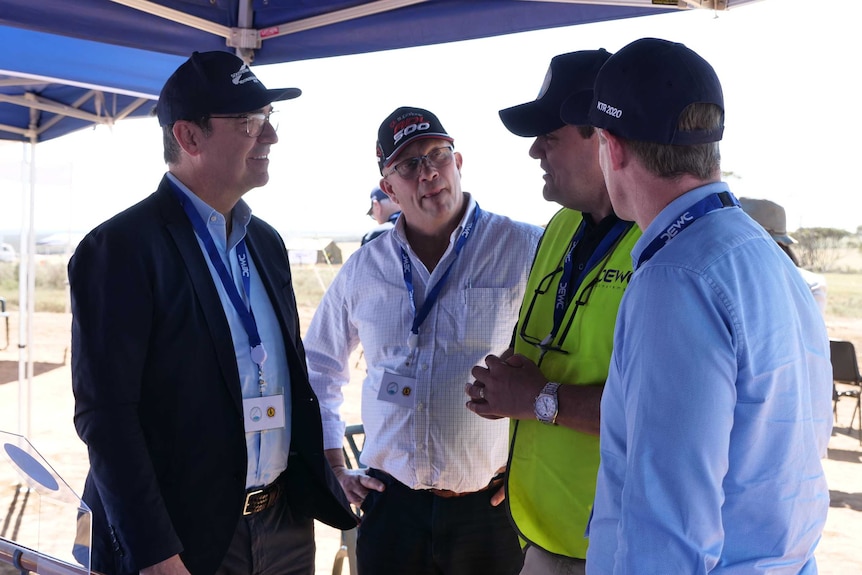 A man wearing a jacket and baseball cap speaks to other men the same under a canopy