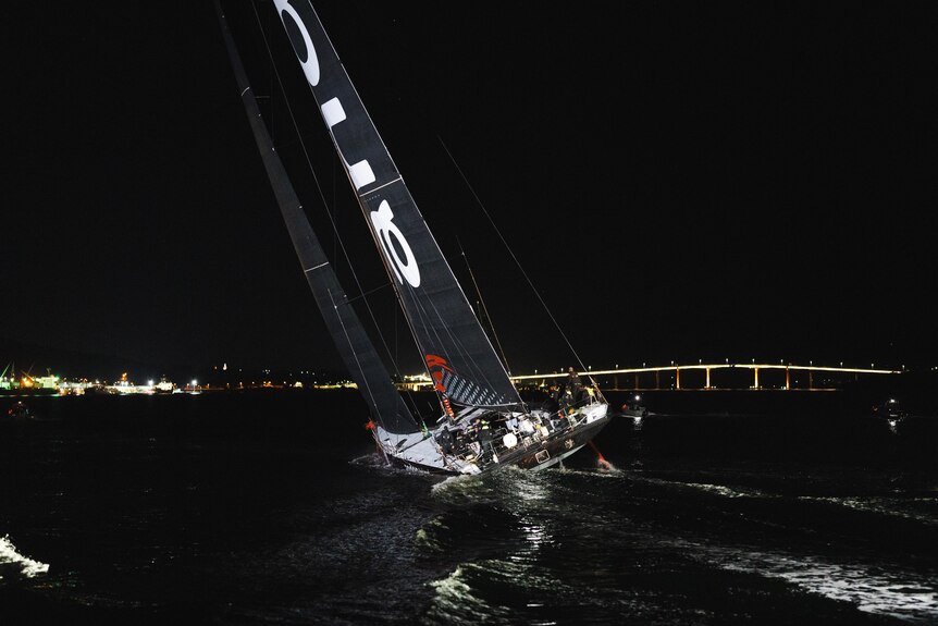 A yacht with a black sail with white writing at night time with a bridge in the background