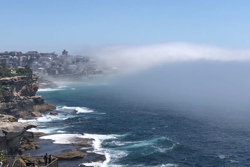 The rocks at Bondi beach are seen to the left of image as sea crashes against them, with fog rising across skyline.