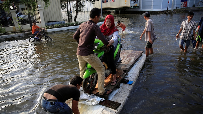 A worker floats her motorcycle on styrofoam to avoid sea water during high tide