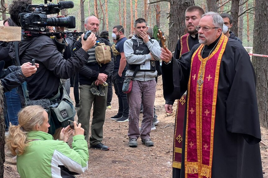 Cameraman filming a priest in robes holding a cross in the street.