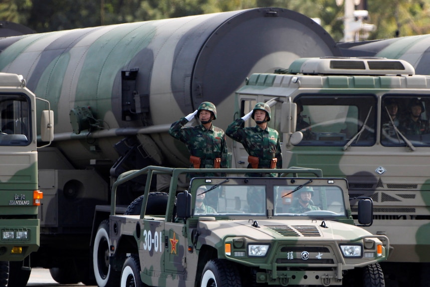 Two soldiers hold their hands up in a salute while they ride in an open truck pulling a giant missile