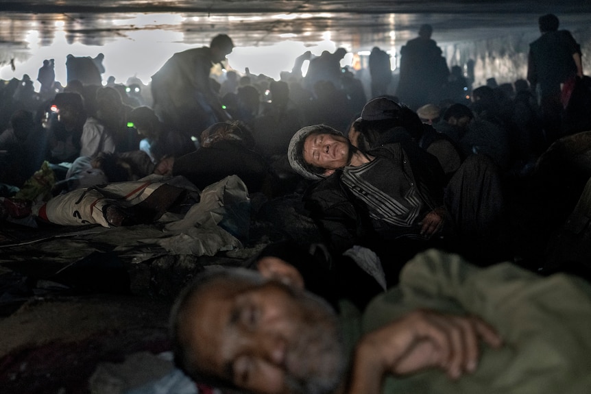 A mass of impoverished-looking people crouch, crawl and lie under a bridge, with daylight visible in the background.