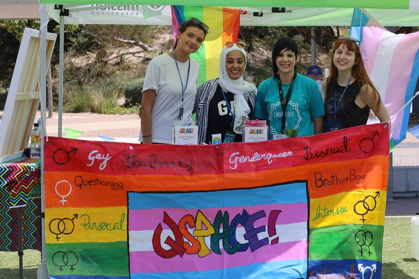 Four young women stand before rainbow sign