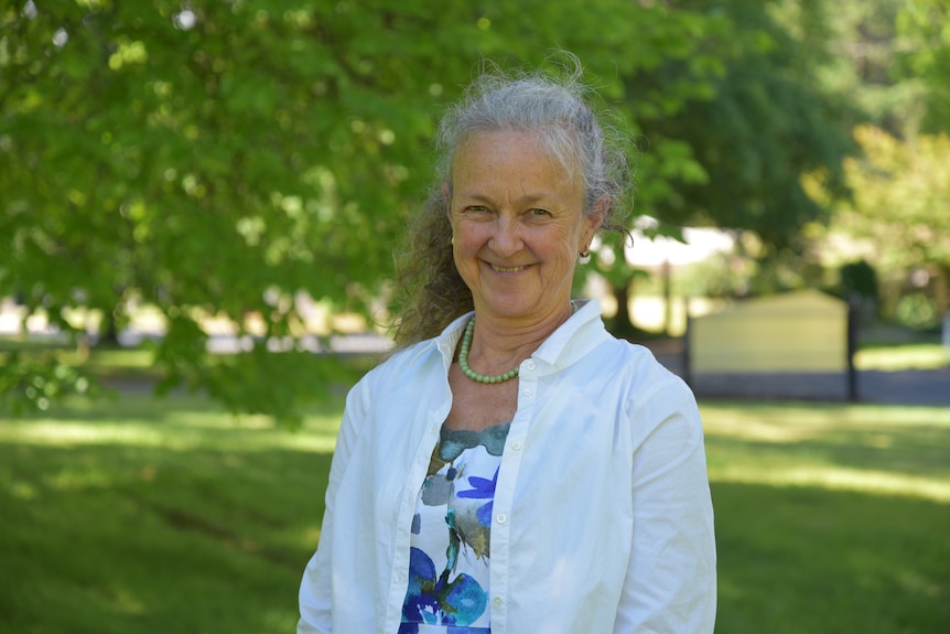 A smiling woman with grey hair wearing a white shirt stands in a green park