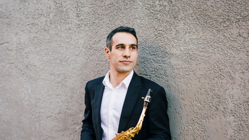 Nick Russoniello holding a saxophone, wearing a suit and standing against a wall.