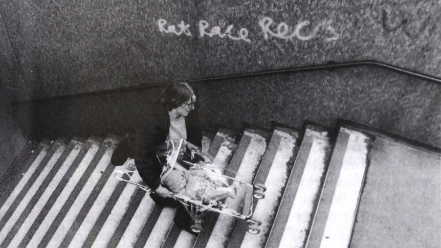 A black and white photo shows a woman carrying a pram up London underground stairs.