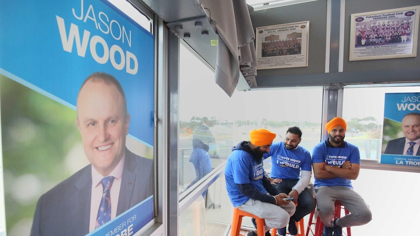 Three men sit in the corner of a room wearing Jason Wood campaign tops. His campaign poster covers a window