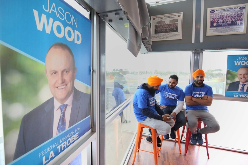 Three men sit in the corner of a room wearing Jason Wood campaign tops. His campaign poster covers a window