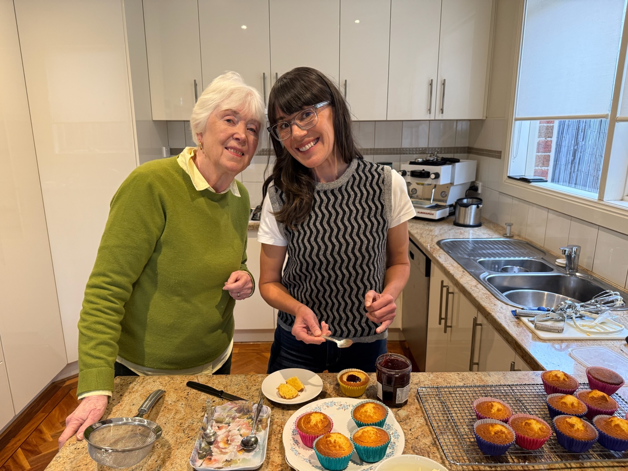 A woman with grey hair stands next to her adult daughter in a kitchen, baking ingredients in front of them on bench