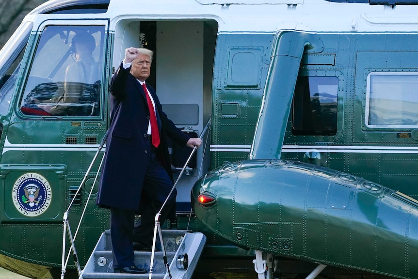 A man wearing a suit with a red tie stands on the steps of a helicopter with his fist in the air