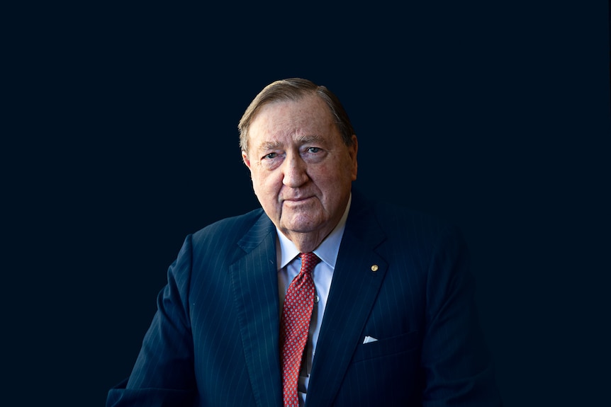 An older man in a suit looks seriously into the camera against a black background.