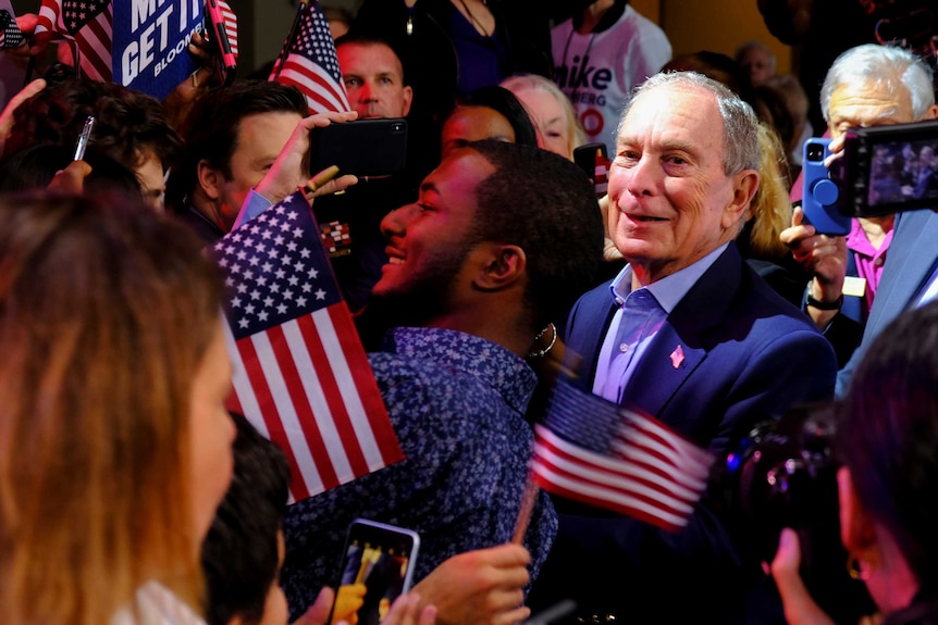 Michael Bloomberg smiling surrounded by Americans flags and fans