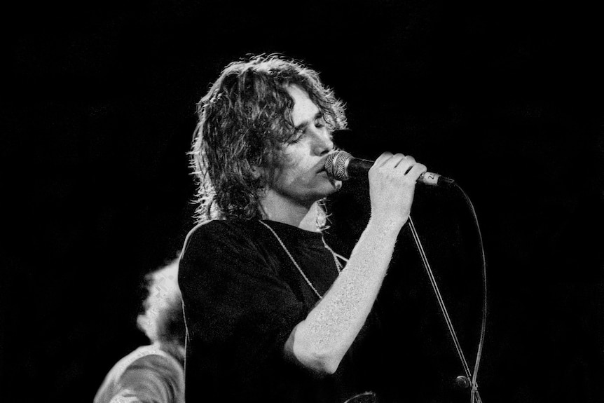 A young man with shaggy hair wearing a black sweater sings into a microphone, his eyes closed