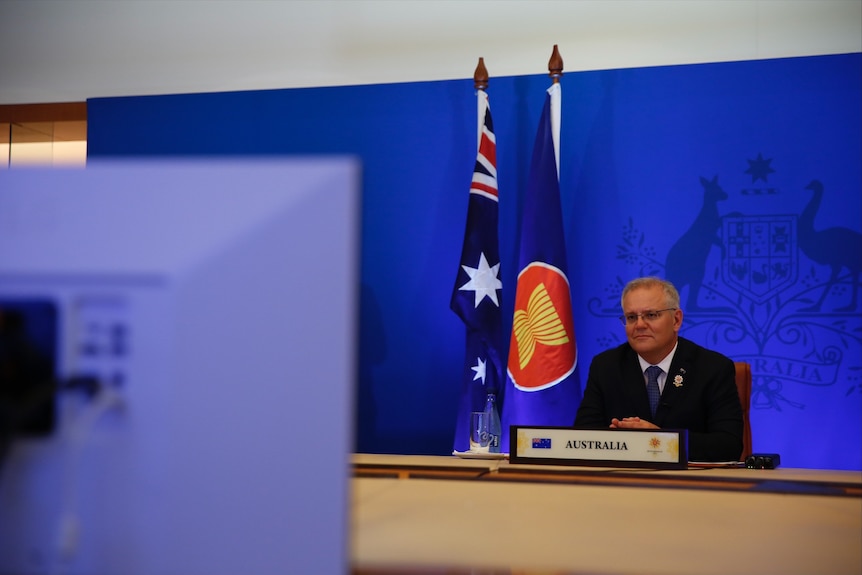 A man in a suit smiles as hit sits behind a desk and looks at a video screen on which a virtual conference is being held.