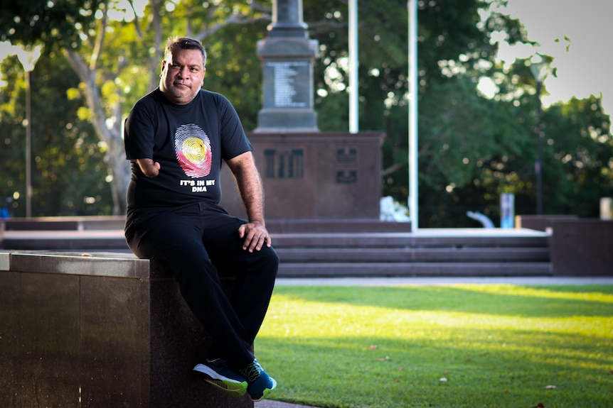 A man wearing a t-shirt with Aboriginal flag colours, sitting on a marble step as part of a war memorial.