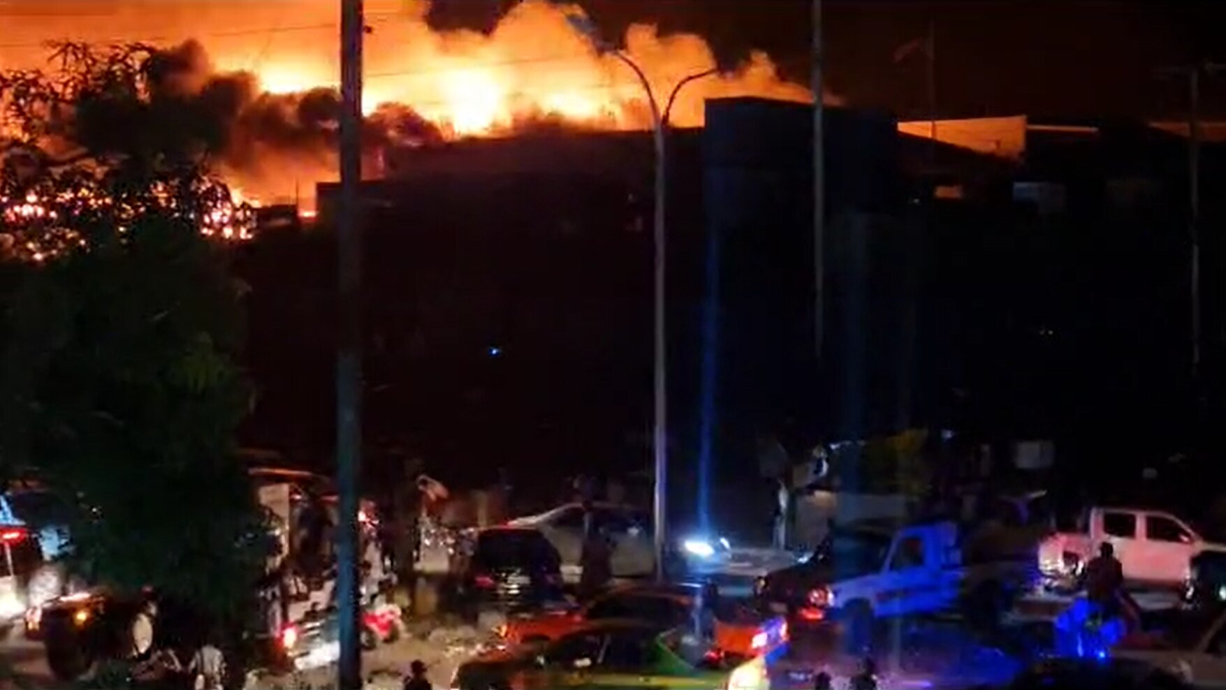 A large building burns as people walk through the street in the night in front of it.