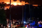 A large building burns as people walk through the street in the night in front of it.